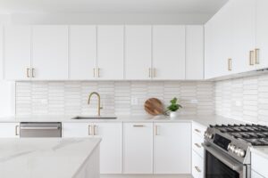 Home 13 A beautiful kitchen detail with white cabinets, a gold faucet, white marble countertops, and a brown picket ceramic tile backsplash.