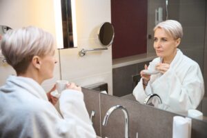 Older woman with tea or coffee looking at herself in mirror in bathroom