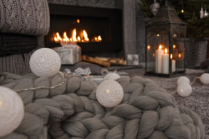 Beautiful winter composition in gray shades with a fireplace, cozy knitted blanket, candles, Christmas gifts and a garland.