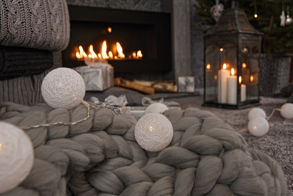 Beautiful winter composition in gray shades with a fireplace, cozy knitted blanket, candles, Christmas gifts and a garland.
